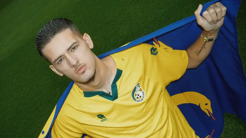 Justin Hubner mengenakan jersey kuning Fortuna Sittard sambil membentangkan bendera klub di atas rumput stadion.